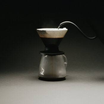 A black Hario V60 Neo with the goose neck of a Hario kettle pouring hot water in to the dripper